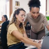 Im Hintergrund arbeiten mehrere Menschen an ihrem Schreibtisch. Vorne sieht man zwei junge Frauen, die gemeinsam auf einen Laptopbildschirm schauen und sich angeregt unterhalten. 