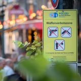 Im Hintergrund ist verschwommen ein Abschnitt der Düsseldorfer Altstadt zu sehen. Davor steht ein gelbes Schild mit der Aufschrift "Waffenverbotszone". Auf dem Schild sind Bilder verschiedener Waffen zu sehen, die mit einem Roten X durchgestrichen sind.